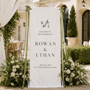 May include: A tall, rectangular white fabric wedding welcome sign featuring black text that reads: R E, WELCOME TO THE WEDDING OF, ROWAN & ETHAN, 25th July, 2025, IT'S A FOREVER KIND OF THING. The banner is suspended by olive green ribbons.
