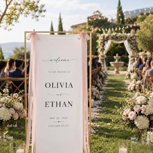 May include: A tall, rectangular gold metal frame holding a white fabric banner with pink ribbon accents. The text reads: welcome TO THE WEDDING OF OLIVIA and ETHAN MAY 24 . 2026 TUSCANY.ITALY. The banner is displayed vertically in an outdoor setting.