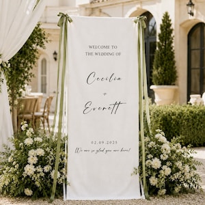 May include: A tall, rectangular white fabric wedding welcome sign featuring black text that reads Welcome to the wedding of Cecilia + Everett, the date 02.09.2025, and We are so glad you are here. The banner is tied with olive green ribbons.