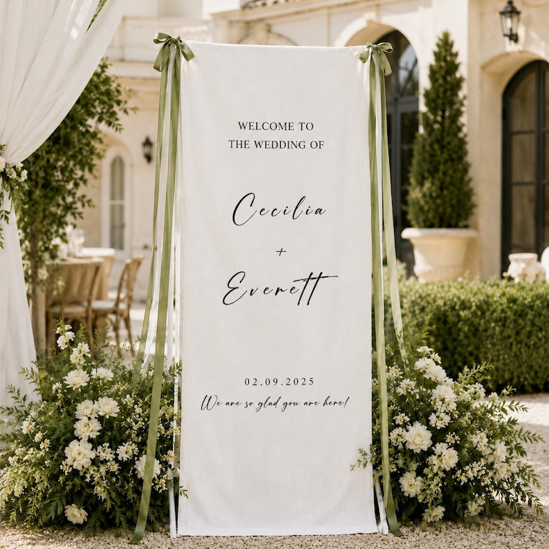 May include: A tall, rectangular white fabric wedding welcome sign featuring black text that reads Welcome to the wedding of Cecilia + Everett, the date 02.09.2025, and We are so glad you are here. The banner is tied with olive green ribbons.