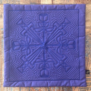May include: A purple square throw pillow with a quilted design featuring a repeating pattern of stylized roses and leaves. The quilting is done in a dark blue thread.
