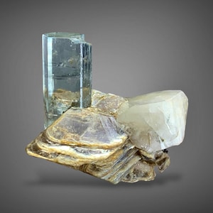 Puede incluir: Un espécimen mineral con una formación cristalina de aguamarina azul claro translúcida. Los cristales de aguamarina son verticales y rectangulares, sobre una base de mica en capas de color marrón claro y un cristal de cuarzo blanco.