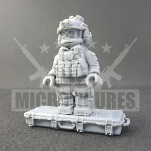 May include: A gray miniature figurine of a soldier wearing a helmet and tactical vest, standing on a gray case. The figurine has a skull-like face. The word "MICRO" is visible on the case.