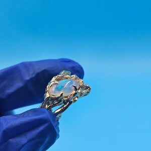 May include: A gold ring featuring an oval opal gemstone with a blue and white iridescent sheen. The ring has a textured, organic design with small accent stones. The ring is held against a bright blue background.
