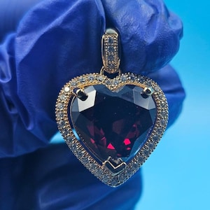 May include: A heart-shaped pendant with a large, faceted, deep red gemstone. The gemstone is surrounded by a gold-colored frame with small, clear accent stones. The pendant is suspended from a gold-colored bail.
