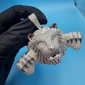 May include: A silver-colored lion pendant encrusted with sparkling, clear gemstones. The pendant features a detailed lion's head with open mouth and sharp teeth, and is held by a black gloved hand against a blue background.