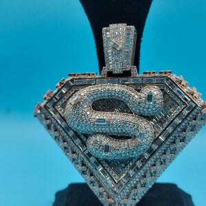 May include: A diamond-encrusted pendant in a shield shape, featuring a stylized "S" in the center. The pendant has a rose gold tone and is set with numerous small diamonds and larger baguette-cut stones.