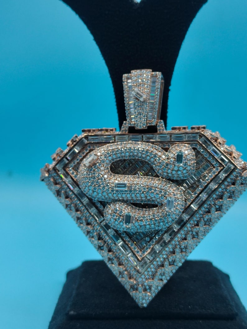 May include: A diamond-encrusted pendant in a shield shape, featuring a stylized "S" in the center. The pendant has a rose gold tone and is set with numerous small diamonds and larger baguette-cut stones.
