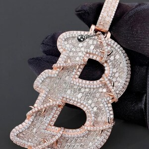 May include: A rose gold pendant in the shape of the letter B, fully encrusted with numerous small, clear baguette and round cut stones. The letter is wrapped in a rose gold vine-like design with small thorns. The text Worldmine is visible.