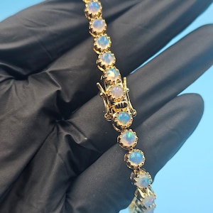 Puede incluir: Una pulsera dorada con una serie de piedras de ópalo redondas e iridiscentes. Los ópalos están engastados en un engaste de bisel dorado y conectados por una cadena dorada. La pulsera se muestra sobre un fondo azul.