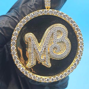 May include: Gold-toned pendant with a black circular center, featuring the letters "MB" in a sparkling, diamond-like design. The pendant is encircled by a row of clear stones and hangs from a matching bail.
