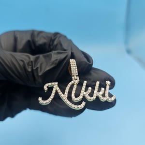 Könnte beinhalten: Ein goldfarbener Anhänger mit dem Namen "Nikki" in einer kursiven Schriftart, besetzt mit kleinen, klaren Edelsteinen. Der Anhänger wird vor einem blauen Hintergrund gehalten.