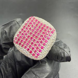 Puede incluir: Un anillo cuadrado con una banda plateada y una superficie cubierta de pequeñas y vibrantes piedras preciosas rosas. Los bordes están bordeados con piedras preciosas claras más pequeñas. El anillo es sostenido por una mano enguantada sobre un fondo oscuro.