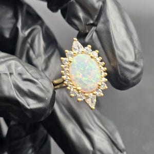 May include: A gold ring featuring an oval opal gemstone surrounded by a halo of small, clear gemstones. The ring's design includes a delicate band and a cluster of pear-shaped and round stones around the opal, creating a sparkling effect.