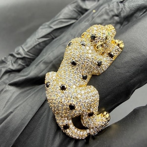 May include: A gold-toned, bejeweled leopard figurine. The leopard is covered in small, clear crystals with black spots and green eyes. The figurine is lying down with its paws crossed.