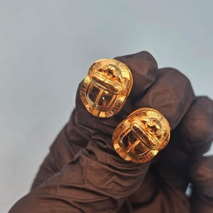 May include: A pair of gold-colored scarab beetle earrings. The earrings are round with a detailed beetle design. The earrings are held by a person wearing a black glove.