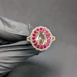 Puede incluir: Un anillo de oro rosa con una gran piedra preciosa ovalada de color melocotón rodeada por un halo de pequeñas piedras rojas y un segundo halo de pequeñas piedras transparentes. El anillo tiene un diseño de caña dividida.