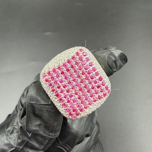 May include: A square ring with a silver band, featuring a surface densely packed with small, bright pink gemstones. The ring is displayed against a dark backdrop, highlighting its sparkle.