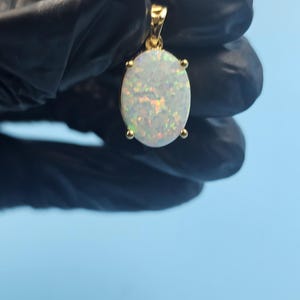 Puede incluir: Un colgante de oro con una piedra preciosa de ópalo ovalada. El ópalo tiene una base blanca con destellos iridiscentes de color. El colgante está engastado en un bisel de oro con cuatro pequeñas garras que sujetan la piedra. La argolla también es de oro.