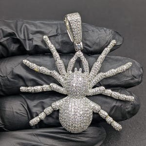 May include: A silver spider pendant, covered in small, glittering stones. The spider design features eight articulated legs and a bail for a chain. The pendant is designed to be worn as jewellery.