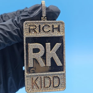 May include: Gold-coloured pendant with the words "RICH" and "KIDD" in a rectangular shape. The letters "RK" are in the centre. The pendant is encrusted with small, sparkling stones. The background is a solid light blue.