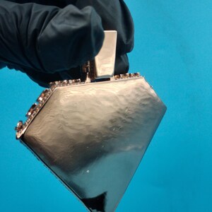 May include: A silver-colored, diamond-shaped pendant with a rectangular top. The pendant is held by a gloved hand against a blue background. The edges of the pendant are lined with small, dark-colored embellishments.