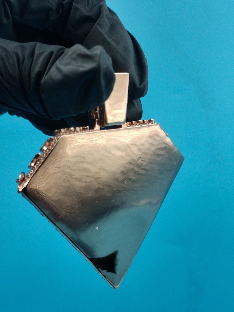 May include: A silver-colored, diamond-shaped pendant with a rectangular top. The pendant is held by a gloved hand against a blue background. The edges of the pendant are lined with small, dark-colored embellishments.