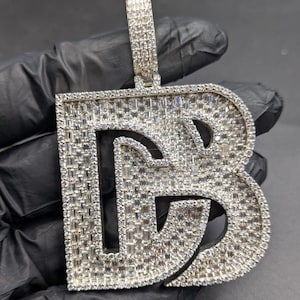 May include: A silver-colored pendant in the shape of the letter "B", encrusted with numerous small, sparkling stones. The pendant has a bail at the top for a chain. The background is a dark, solid color.