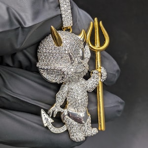 Puede incluir: Un colgante con incrustaciones de diamantes que representa a un diablo de dibujos animados sosteniendo un tridente dorado. El diablo tiene cuernos dorados y un taparrabos plateado. El colgante está sobre un fondo oscuro.
