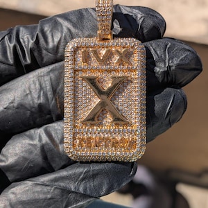 Pode incluir: Pingente retangular dourado com uma moldura cravejada de diamantes. O pingente apresenta o número romano "X" no centro, com "IVX" acima e "KENTROX" abaixo. A argola também é cravejada com diamantes.