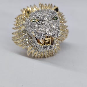May include: A gold and silver lion head ring. The lion's head is encrusted with small, sparkling stones, with green stones for the eyes. The mane is gold, and the mouth is open, revealing gold teeth. The ring is a statement piece.