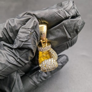 May include: A gold-toned boxing glove pendant featuring a polished finish on the thumb and wrist area, while the main body and cuff are encrusted with numerous small, clear, sparkling stones. A large, smooth gold bail is attached to the top.