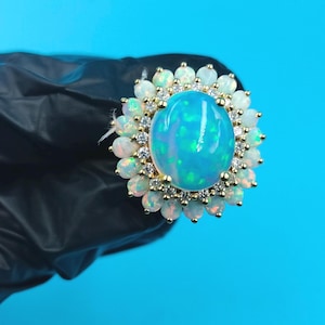 Könnte beinhalten: Ein goldfarbener Ring mit einem großen, ovalen, irisierenden blauen Opal-Edelstein in der Mitte, umgeben von kleineren Opalen und klaren Edelsteinen. Der Ring hat ein Blumenmuster, wobei die Opale in einem Sonnenstrahlenmuster angeordnet sind. Der Ring ist vor einem schwarzen Hintergrund platziert.