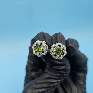 Peut inclure: Une paire de boucles d'oreilles à clous avec une pierre précieuse centrale ovale verte claire, entourée d'un halo de petites pierres précieuses claires. Les boucles d'oreilles ont un design floral et sont serties dans un métal de couleur argent. Le fond est bleu clair uni.