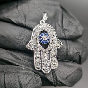 Peut inclure: Pendentif en forme de main de Hamsa argenté avec un motif d'œil bleu au centre. Le pendentif est incrusté de petites pierres précieuses transparentes, et l'œil présente un cercle de pierres bleues. Conçu comme bijou.