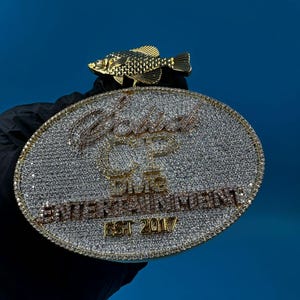 May include: A gold-colored pendant with an oval shape, encrusted with small, sparkling stones. The pendant features the text "Gold CP Entertainment EST 2017" and is topped with a detailed gold fish.
