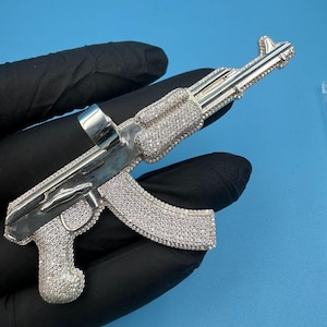 以下が含まれることがあります： 小さな輝く石がちりばめられた、AK-47ライフル型のシルバーペンダント。 青い背景に、黒い手袋をはめた手でペンダントが持たれています。 デザインには、銃器の詳細な特徴が含まれています。