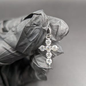 Peut inclure: Un pendentif en argent en forme de croix orné de cinq pierres précieuses rondes et transparentes. La croix est présentée sur un fond sombre. Les pierres sont disposées en croix classique, reflétant la lumière.