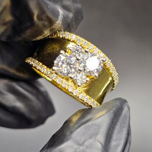 Peut inclure: Une bague en or avec une large bande, ornée d'un groupe de pierres précieuses claires au centre et de petites pierres sur les bords. La bague est tenue sur un fond sombre.