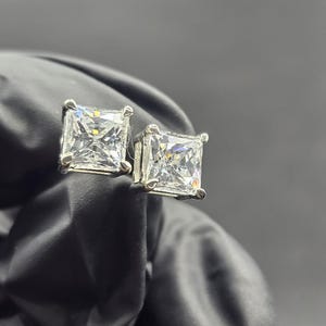 Peut inclure: Une paire de boucles d'oreilles à clous carrées en pierres précieuses transparentes. Les pierres sont serties dans un métal argenté. Les boucles d'oreilles sont présentées sur un fond sombre, soulignant leur éclat et leur clarté.