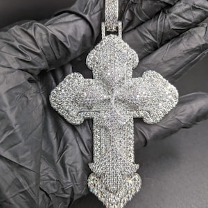May include: A silver-colored cross pendant encrusted with numerous small, sparkling stones. The cross has a detailed design with rounded edges and a decorative bail for a chain. The pendant is held against a dark background.