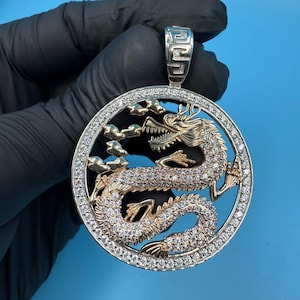 Puede incluir: Colgante redondo con un diseño de dragón dorado finamente detallado, enmarcado en plata. El dragón está adornado con piedras brillantes, y el marco está adornado con piedras más pequeñas. El colgante tiene una fianza plateada.