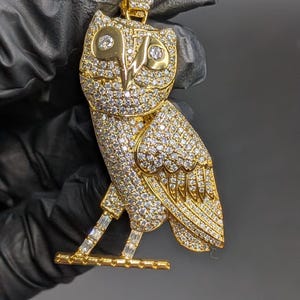 May include: A gold-colored owl pendant encrusted with numerous small, sparkling gemstones. The pendant features an owl design with detailed wings, eyes, and a perch. The owl is set against a dark background.