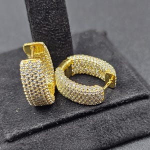 Peut inclure: Une paire de boucles d'oreilles créoles dorées incrustées de petits pierres précieuses transparentes. Les boucles d'oreilles sont exposées sur une surface noire, mettant en valeur leurs détails scintillants. Les boucles d'oreilles sont de forme rectangulaire.