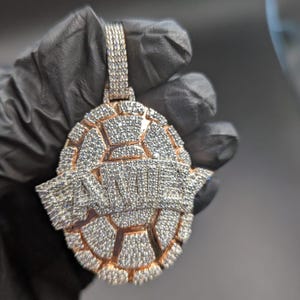 Op de afbeelding: Een grote, sierlijke hanger met een geometrisch ontwerp, ingelegd met talrijke kleine, sprankelende stenen. De hanger heeft een goudkleurige rand en een beugel aan de bovenkant voor een ketting. Het woord "ANIMALS" staat in het midden van de hanger.