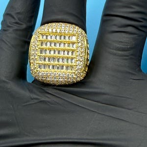 Puede incluir: Un anillo dorado con una cara cuadrada, incrustado con numerosas piedras preciosas pequeñas y transparentes. El centro presenta un patrón de rejilla de piedras rectangulares. El anillo está sobre un fondo negro.