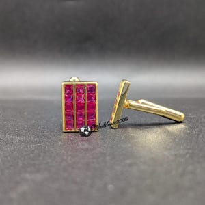 May include: A pair of gold-tone cufflinks featuring a rectangular face with a grid of nine square-cut, vibrant pink stones. One cufflink is shown from the front, while the other is positioned to display the side profile and the metal toggle mechanism.
