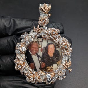 May include: A circular pendant featuring a central photograph of two people framed by rose gold and silver-toned metal flowers and hummingbirds. The entire frame is encrusted with clear, sparkling stones, and a hummingbird sits atop the bail.