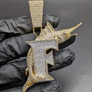 Può includere: Un ciondolo dorato a forma di pesce marlin, incastonato con scintillanti pietre preziose bianche e gialle. Il ciondolo presenta la lettera "F" in un design stilizzato. Il pesce ha un muso lungo e appuntito e pinne dettagliate.