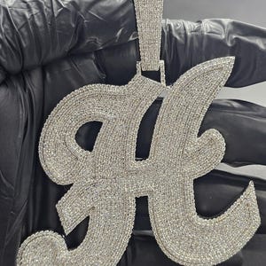 May include: A large, silver pendant shaped like the letter "H", covered in small, clear gemstones. The pendant has a brilliant, sparkling appearance. The bail, used for attaching to a chain, is also adorned with gemstones.